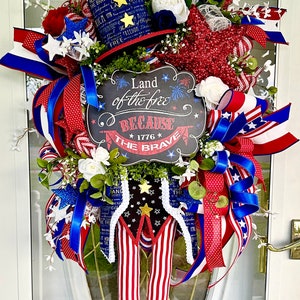 May include: A red, white, and blue patriotic wreath with a chalkboard sign that reads "Land of the free because 1776 the brave". The wreath is decorated with stars, ribbons, and flowers.