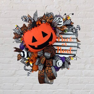 Puede incluir: Una corona de Halloween con una cinta a rayas blancas y negras, malla naranja y negra y una calabaza de peluche con una cara de jack-o'-lantern. La corona tambi&eacute;n tiene un letrero que dice "Trick or Treat".