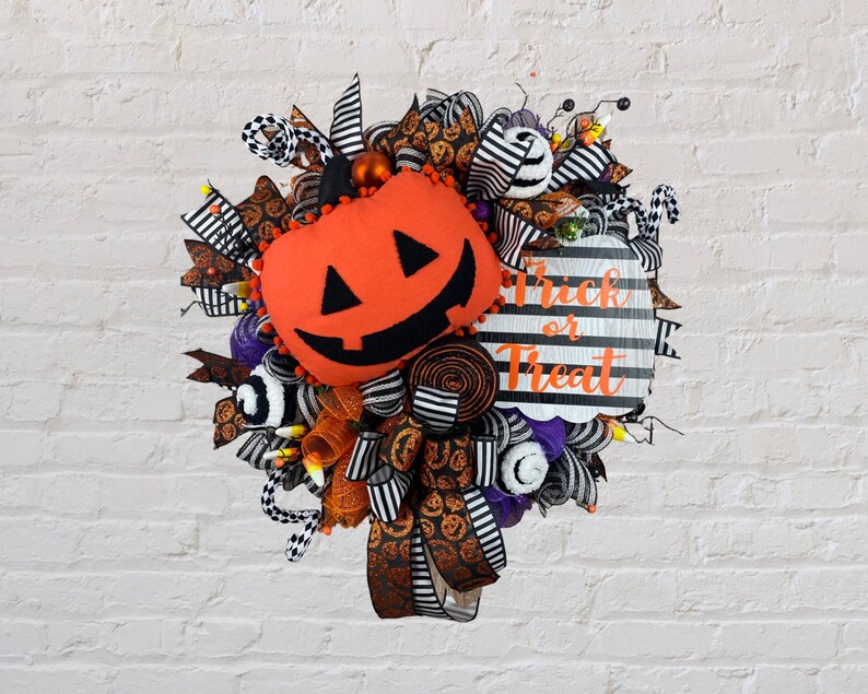 Puede incluir: Una corona de Halloween con una cinta a rayas blancas y negras, malla naranja y negra y una calabaza de peluche con una cara de jack-o'-lantern. La corona tambi&eacute;n tiene un letrero que dice "Trick or Treat".