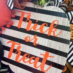 Puede incluir: Un letrero con forma de calabaza a rayas blancas y negras con las palabras "Trick or Treat" en letras naranjas. El letrero est&aacute; decorado con una ara&ntilde;a brillante.