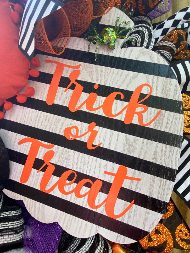 Puede incluir: Un letrero con forma de calabaza a rayas blancas y negras con las palabras "Trick or Treat" en letras naranjas. El letrero est&aacute; decorado con una ara&ntilde;a brillante.