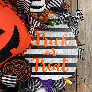 Puede incluir: Una corona de calabaza a rayas blancas y negras con las palabras "Trick or Treat" en letras naranjas. La corona est&aacute; decorada con cinta a rayas blancas y negras, pompones blancos y negros, purpurina naranja y negra y un bast&oacute;n de caramelo blanco y negro.
