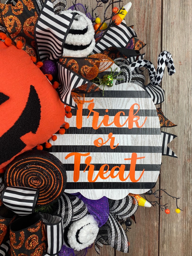 Puede incluir: Una corona de calabaza a rayas blancas y negras con las palabras "Trick or Treat" en letras naranjas. La corona est&aacute; decorada con cinta a rayas blancas y negras, pompones blancos y negros, purpurina naranja y negra y un bast&oacute;n de caramelo blanco y negro.