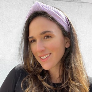 May include: A woman with long brown hair is wearing a light purple knotted headband. She is wearing a black long-sleeved shirt.