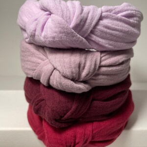 May include: Four fabric headbands stacked on top of each other. The top headband is light purple, the second is a darker shade of purple, the third is burgundy, and the bottom headband is a deep red.