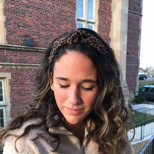 May include: A woman with long, curly brown hair is wearing a light brown fluffy sweater and a leopard print headband. She is standing in front of a brick building.