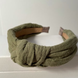 May include: A green fabric headband with a knotted design. The headband is made of a soft, textured fabric and has a simple, elegant style.