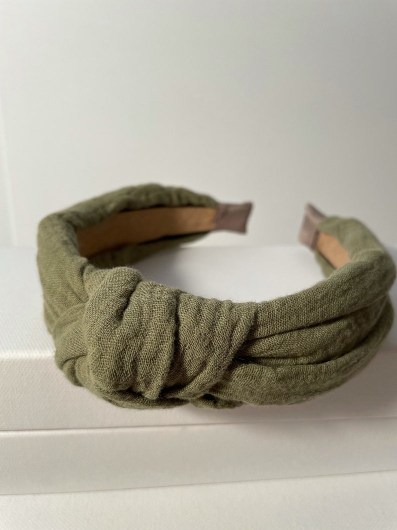 May include: A green fabric headband with a knotted design. The headband is made of a soft, textured fabric and has a simple, elegant style.