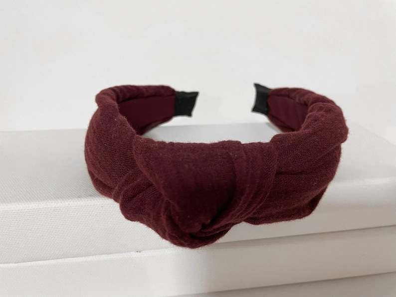 May include: A burgundy knotted headband made of fabric. The headband is a simple design with a single knot in the center.