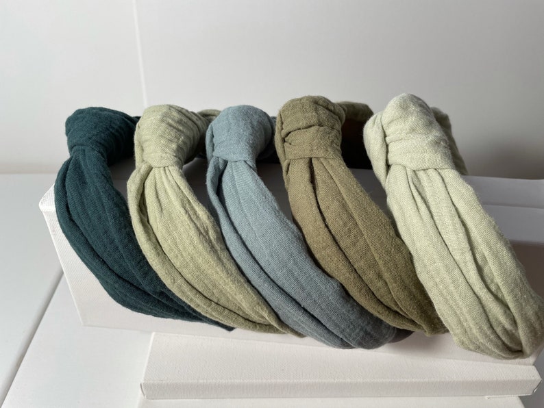 May include: Five fabric headbands in different shades of green, blue, and gray. The headbands are made of a soft, textured fabric and have a knotted design.