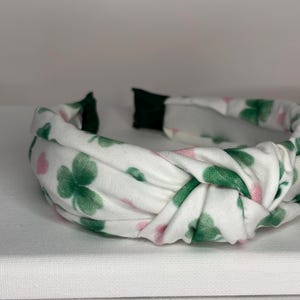 K&ouml;nnte beinhalten: Ein wei&szlig;es Stirnband mit einem Muster aus gr&uuml;nen Kleebl&auml;ttern und rosa Blumen. Das Stirnband ist verknotet und hat ein elastisches Gummiband.