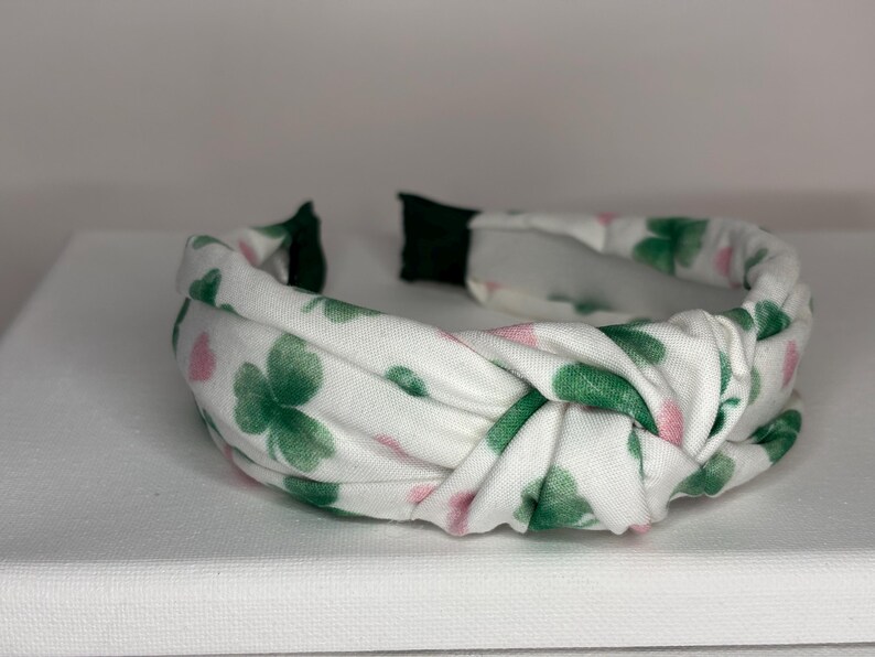 K&ouml;nnte beinhalten: Ein wei&szlig;es Stirnband mit einem Muster aus gr&uuml;nen Kleebl&auml;ttern und rosa Blumen. Das Stirnband ist verknotet und hat ein elastisches Gummiband.