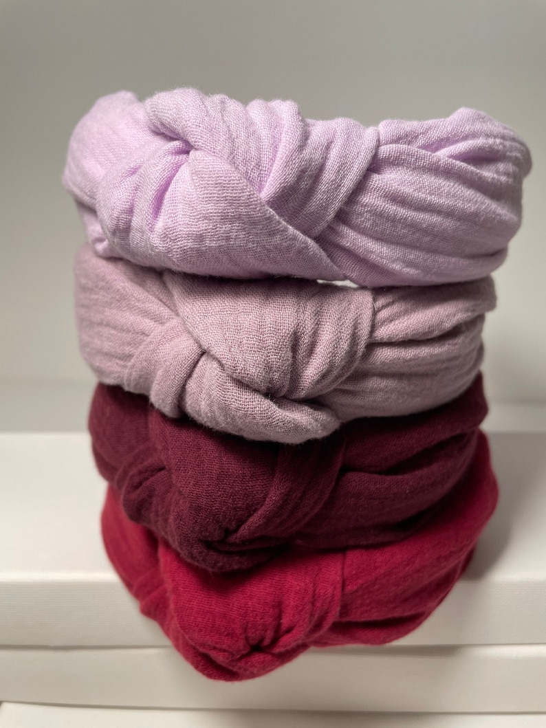 May include: Four fabric headbands stacked on top of each other. The top headband is light purple, the second is a darker shade of purple, the third is burgundy, and the bottom headband is a deep red.