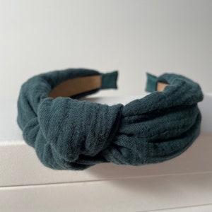 May include: A dark green fabric headband with a knotted design. The headband is made of a soft, textured fabric.