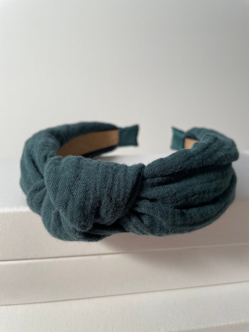 May include: A dark green fabric headband with a knotted design. The headband is made of a soft, textured fabric.