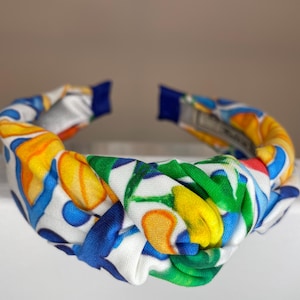 May include: A white knotted headband with a colorful floral pattern featuring yellow, blue, green, and red accents.