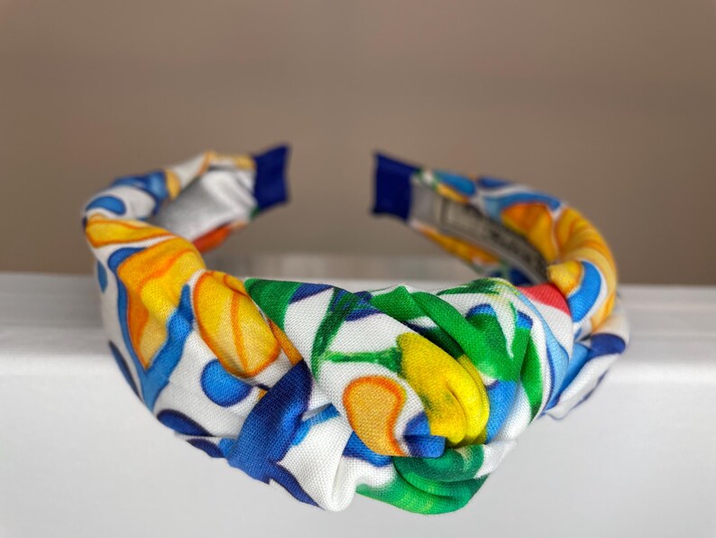 May include: A white knotted headband with a colorful floral pattern featuring yellow, blue, green, and red accents.