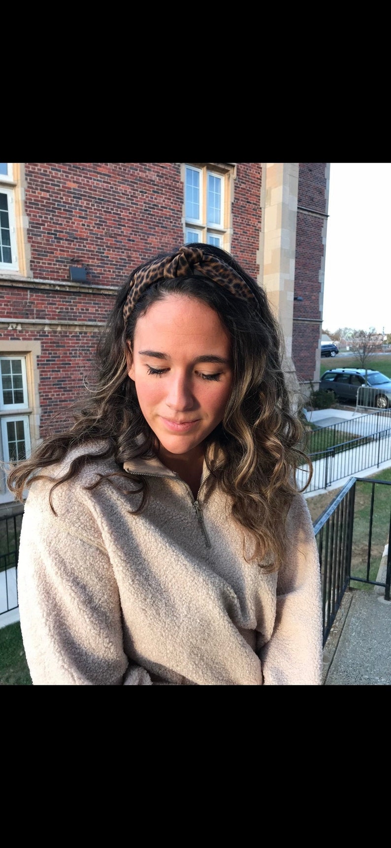 May include: A woman with long, curly brown hair is wearing a light brown fluffy sweater and a leopard print headband. She is standing in front of a brick building.
