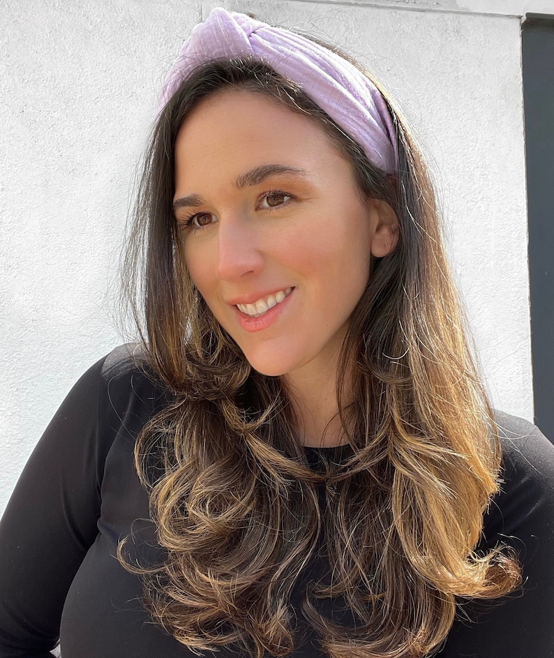 May include: A woman with long brown hair is wearing a light purple knotted headband. She is wearing a black long-sleeved shirt.