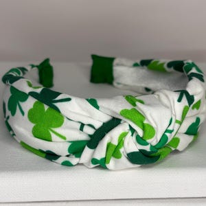 K&ouml;nnte beinhalten: Ein wei&szlig;es Stirnband mit einem gr&uuml;nen Kleeblatt-Muster. Das Stirnband ist geknotet und hat eine weiche, dehnbare Haptik.