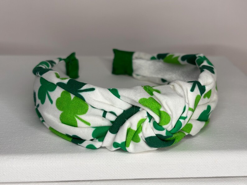 K&ouml;nnte beinhalten: Ein wei&szlig;es Stirnband mit einem gr&uuml;nen Kleeblatt-Muster. Das Stirnband ist geknotet und hat eine weiche, dehnbare Haptik.