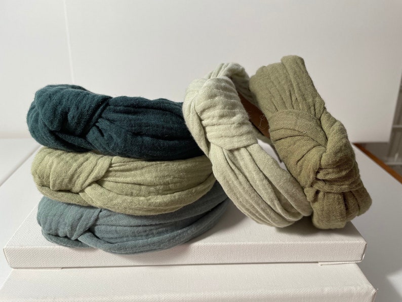 May include: Four fabric headbands in different colors: dark green, light green, blue-green, and beige. The headbands are knotted and have a simple, elegant design.