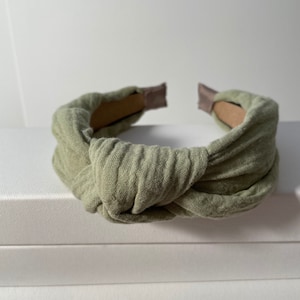 May include: A sage green knotted headband made of soft fabric. The headband is designed to be worn around the head and has a simple, elegant style.