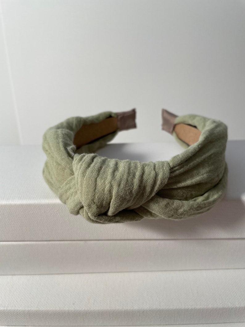 May include: A sage green knotted headband made of soft fabric. The headband is designed to be worn around the head and has a simple, elegant style.