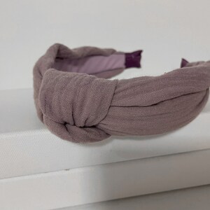 May include: A mauve fabric headband with a knotted design. The headband is made of a soft, textured fabric.
