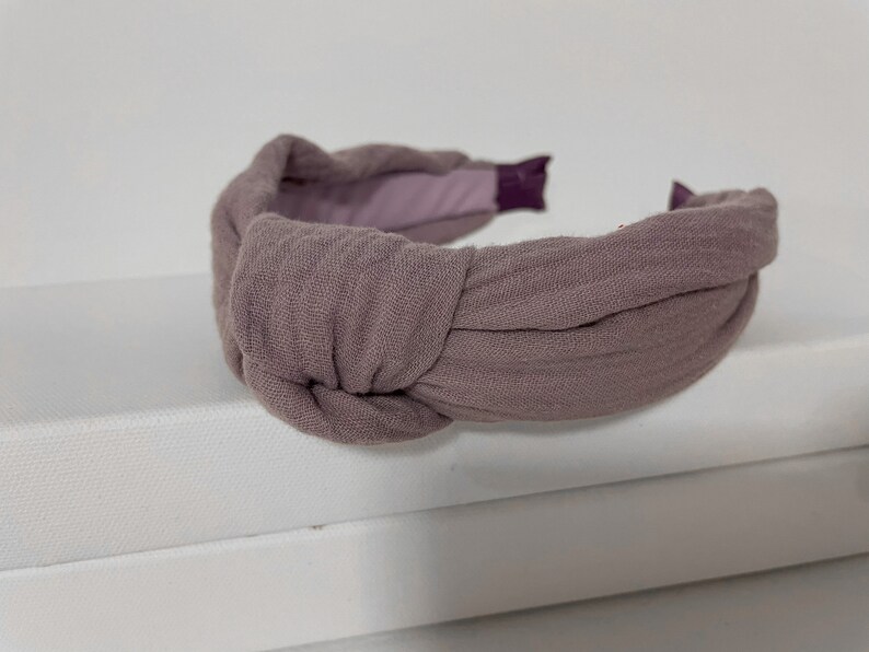 May include: A mauve fabric headband with a knotted design. The headband is made of a soft, textured fabric.
