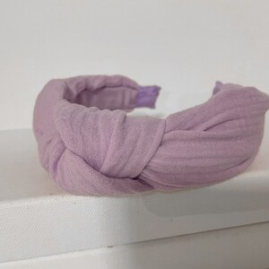 May include: A lavender fabric headband with a knotted design. The headband is made of a soft, textured fabric.