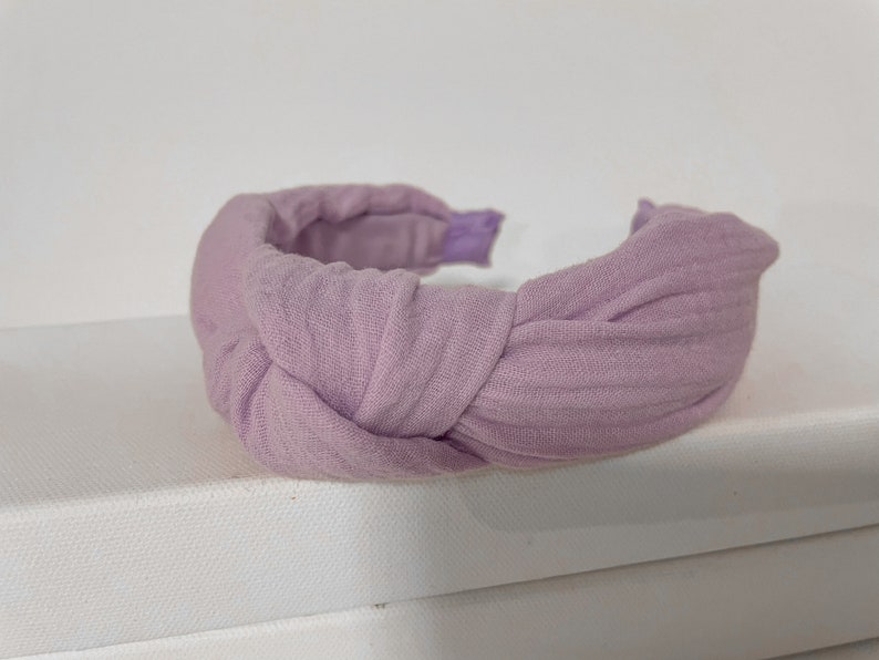 May include: A lavender fabric headband with a knotted design. The headband is made of a soft, textured fabric.