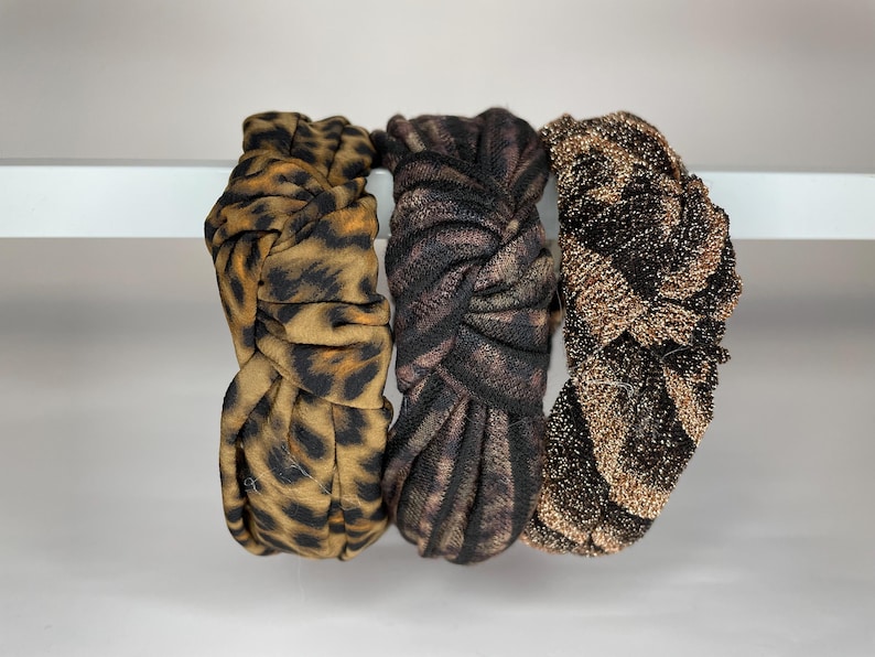 May include: Three fabric headbands in a knotted style. One headband is brown and black with a leopard print, one is black and brown with a swirl pattern, and one is gold and black with a sparkly finish.