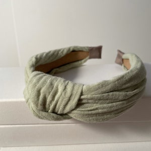 May include: A light green fabric headband with a knotted design. The headband is made of a soft, textured fabric and has a simple, elegant style.