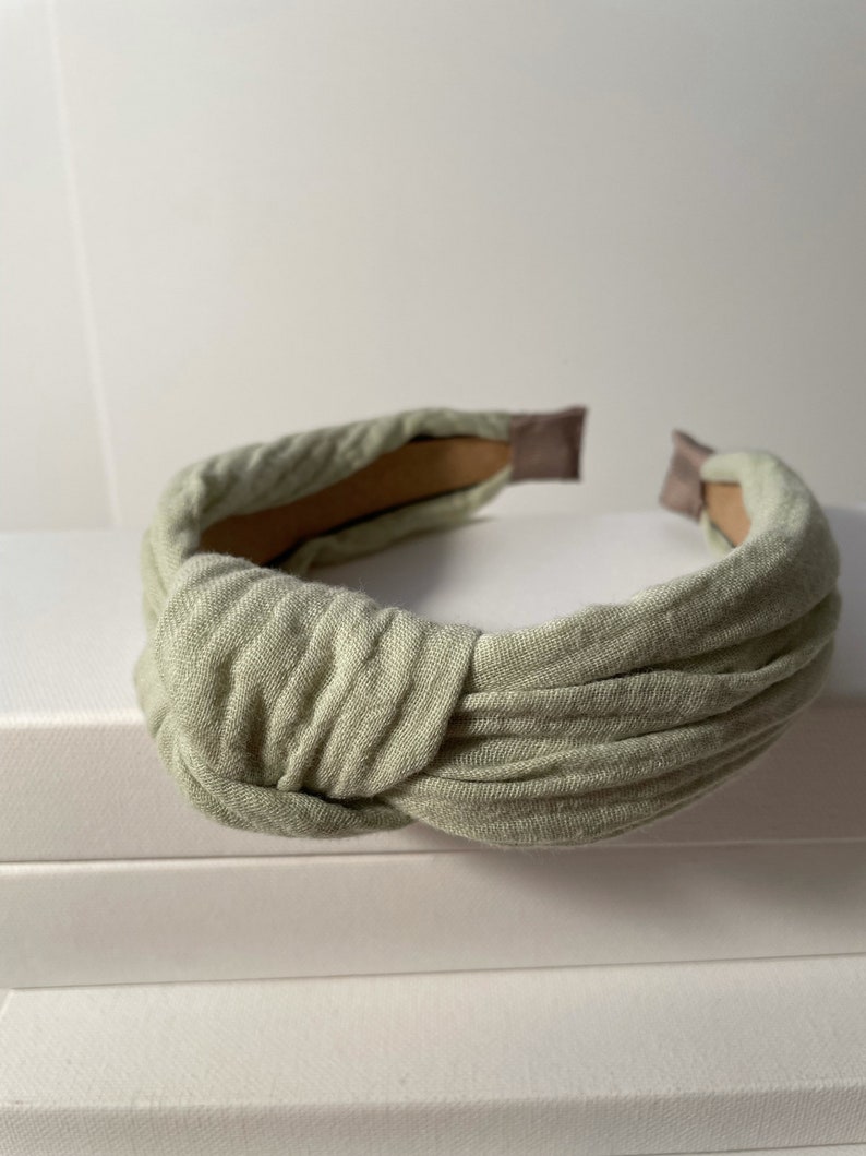 May include: A light green fabric headband with a knotted design. The headband is made of a soft, textured fabric and has a simple, elegant style.