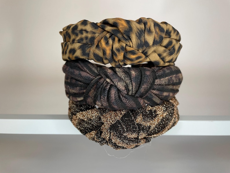 May include: Three knotted headbands stacked on top of each other. The top headband is brown and black with a leopard print. The middle headband is black and brown with a zebra print. The bottom headband is gold with a sparkly finish.