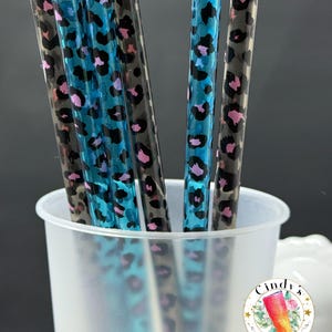 May include: A collection of decorative straws with a leopard print design in shades of black, blue, and pink. The straws are displayed in a clear, frosted plastic cup. A small sticker with text is visible.