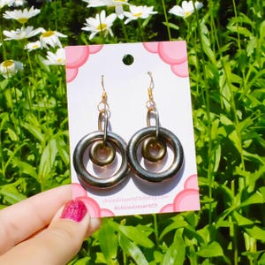 May include: Gold-toned hoop earrings with a smaller inner hoop, displayed on a pink and white card. The card has the text "chloedoesart03@etsy.com" and "@chloedoesart03". The background shows green foliage and white flowers.
