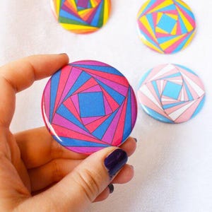 May include: Four colourful round buttons with geometric designs. One button is held in a hand, featuring a blue, pink, and purple pattern. Other buttons display rainbow, pink, yellow, and blue designs. The buttons are on a white surface.