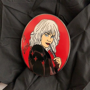 May include: A silver enamel pin featuring a woman with white hair, wearing a black jacket and holding a knife. The background is red with silver glitter stars.