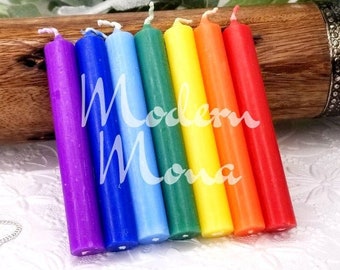 May include: A collection of seven rainbow-colored candles arranged diagonally, with a gemstone pendulum in the foreground. The candles are purple, blue, green, yellow, orange, and red. The text "Modern Mona" is visible on the candles.