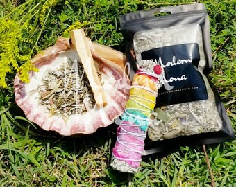 Puede incluir: Un surtido de artículos espirituales sobre hierba verde. Un cuenco de concha contiene hierbas secas y un palo de madera. Un colorido palo de manchado está al lado de una bolsa negra con el texto "Modern Mona" y una dirección web.