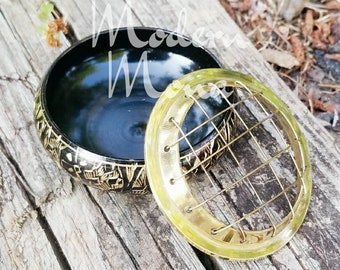 May include: A black bowl with gold etched designs and a removable, clear, circular floral frog. The floral frog has a grid of gold wires. The bowl and frog are on a weathered wooden surface.