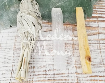 Puede incluir: Un manojo de salvia blanca seca atada con hilo, un cristal de selenita transparente y un palo santo de color marrón claro. Los artículos están dispuestos sobre una superficie de madera blanqueada con una hoja verde en el fondo. El texto "Modern Moms" es parcialmente visible.