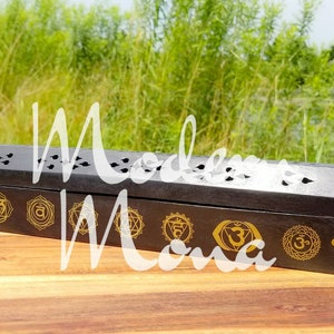 Op de afbeelding: Een zwarte houten wierookhouder met zeven gouden chakra-symbolen. De rechthoekige houder heeft decoratieve uitsparingen en de tekst "Modern Mana". Het object wordt gepresenteerd op een houten oppervlak.