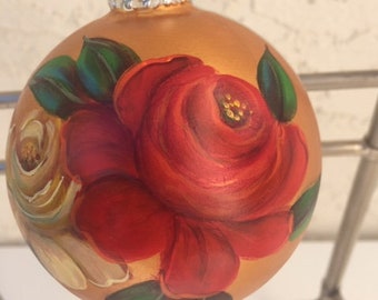 Peut inclure: Une boule de Noël dorée avec un motif peint à la main de roses rouges et blanches. La boule a un sommet argenté et est prête à être accrochée à un arbre.