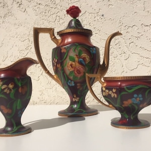 May include: A vintage tea set with a teapot, creamer, and sugar bowl. The set is decorated with a floral pattern in shades of red, green, and blue. The teapot has a red flower on top of the lid.