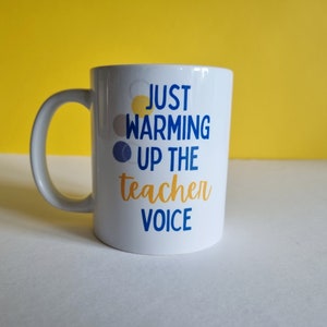 May include: White ceramic mug with a blue and yellow design that says "Just warming up the teacher voice".