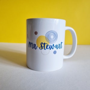 May include: White ceramic mug with a blue and yellow abstract design and the text "mr Stewart" printed on the front.