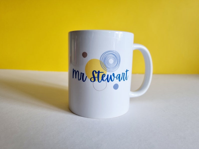 May include: White ceramic mug with a blue and yellow abstract design and the text "mr Stewart" printed on the front.